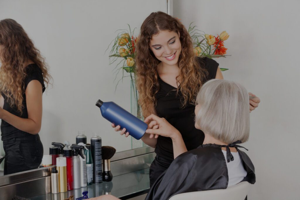 A smiling young hairdresser with curly hair holds a blue haircare bottle while advising a silver-haired older woman seated in a salon chair.