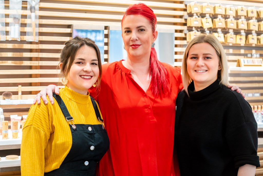 Three smiling female salon professionals pose together with their arms around each other in front of a wooden slatted wall displaying cosmetics. The woman in the centre has bright red hair and a red top.