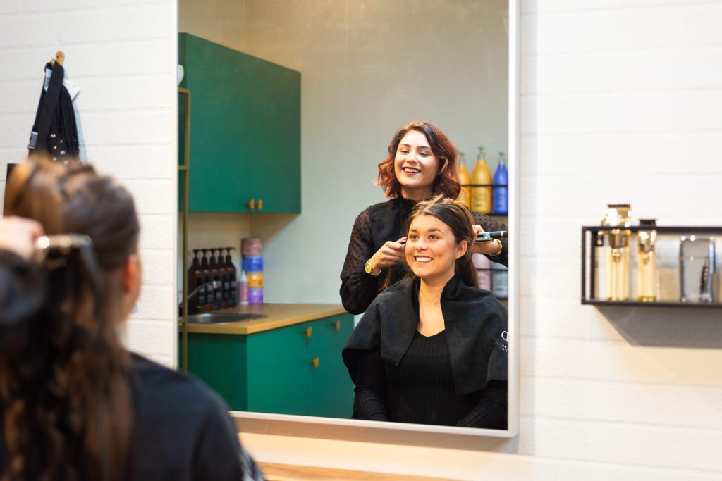 A smiling female hairdresser with auburn hair style's a smiling customer's hair with a straightener, seen through a large rectangular mirror reflection inside a modern salon with emerald green cabinets.