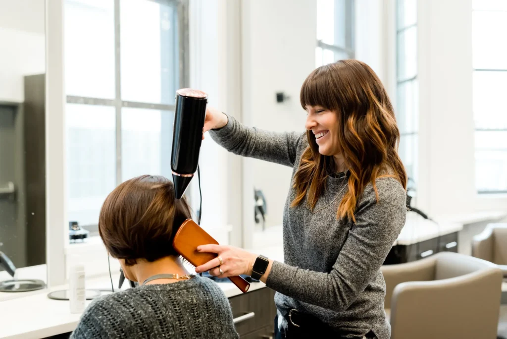 A smiling hairdresser blow-dries and brushes a client's hair in a bright, modern salon.